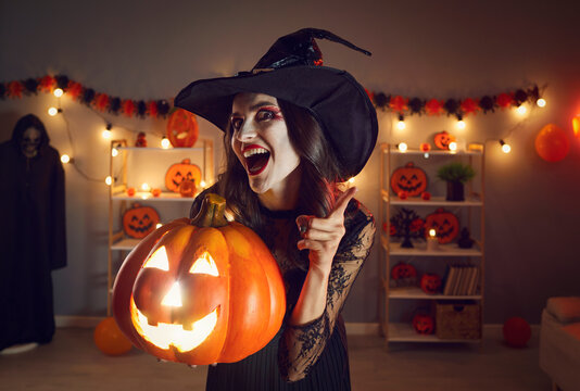 Portrait of woman in wicked witch costume. Beautiful lady in black pointed hat with happy excited face expression standing in dark room, holding Halloween jack-o-lantern pumpkin, shaking finger at you