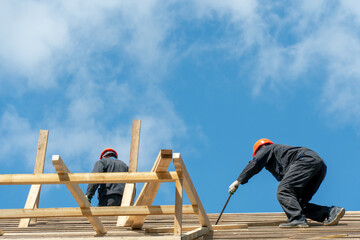 Repair of a wooden roof outdoors on a summer day against the background of blue sky and clouds. A carpenter in special clothes and with a tool installs beams and wood boards.