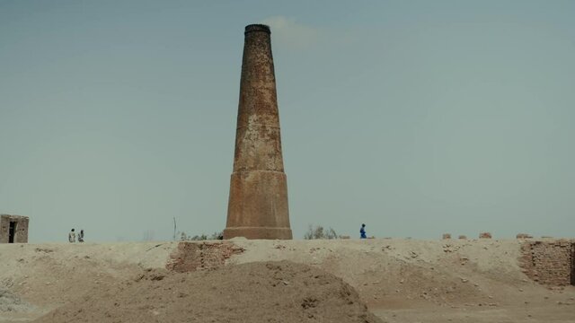 A brick factory in Pakistan that uses slave labor. On the sand there is an empty trailer. Big brick pipe with smoke on background. Slow motion
