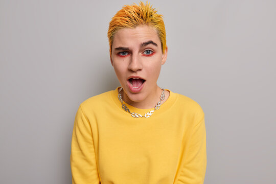 Impressed hipster girl with dyed yellow short hair has bright makeup keeps mouth wide opened feels surprised from hearing shocking news dressed in casual jumper isolated over grey background.