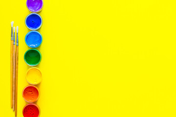 Cans of paint stand in a line on the side, the colors are displayed in the order of the rainbow, there are paint brushes on a yellow background nearby. top view, flat lay, copy space, isolate