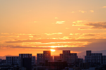 都市の夜明け、早朝ビルの隙間から太陽が昇り辺りはオレンジ色に染まる。ビルはシルエットに浮かぶ。