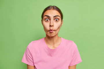 Fototapeta premium Surprised young woman makes fish lips being not scared to show emotions tries to make her crying friend laugh dressed in casual pink t shirt isolated over green background. Face expressions.