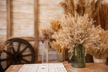 In green glass vase, there was bouquet of dried flowers on wooden bench near backdrop of rural style
