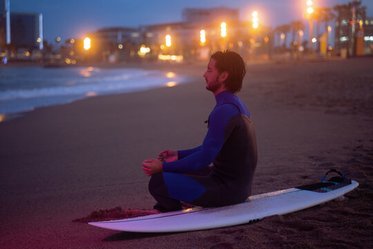 Night portrait of surfer sitting on his surf board and meditating in lotus pose after surfing at Barcelona beach