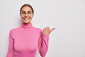 Waist up shot of pretty smiling woman points thumb away advertises your product promots sale or discount dressed in casual pink turtleneck shows free place for design. People and advertisement