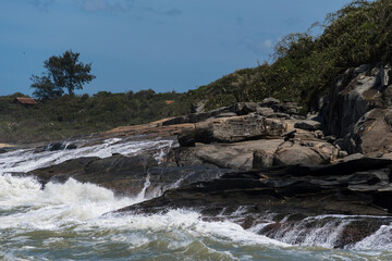 View of Areia Negras beach in Rio das Ostras in Rio de Janeiro with sunny day, blue sky and some clouds. Strong sea and black sand mixed with yellow and many rocks.