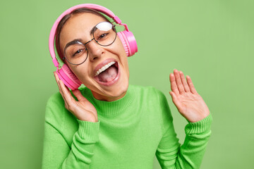 Positive optimistic young European woman laughs happily listens favorite music in pink headphones enjoys audio track wears round transparent glasses and turtleneck isolated over green background.