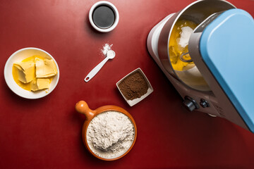 Top view of an electric mixer, flour, butter, cocoa and other ingredients on a red table