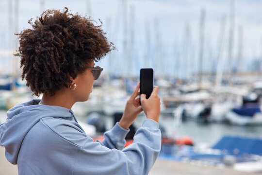 Tourist girl takes photo of yachts in sea port holds mobile phone makes picture during trip inharbor explores destination stands sideways at camera dressed casually. People and vacation concept