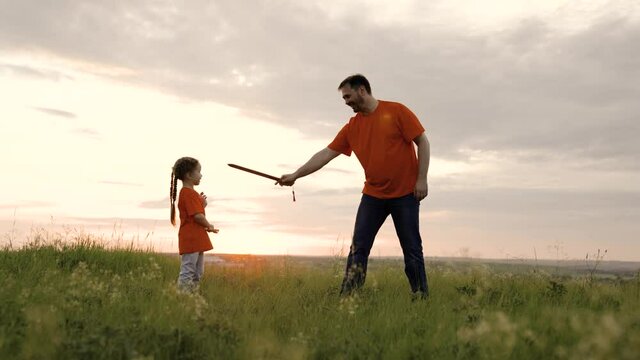Daughter and dad play with toy swords in their hands on the field, playing medieval knights. The child and the father fight with toy sword. Baby and Daddy are playing knights. Happy childhood, family