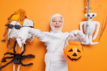 Glad small kid pretends being ghost or mummy holds halloween carved pumpkin stands against orange...