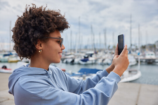 Outdoor shot of curly haired ethnic woman going to have cruise ship vacation enjoys view of sea port makes selfie on modern smartphone wears sunglasses and hoodie takes photo of herself in harbor