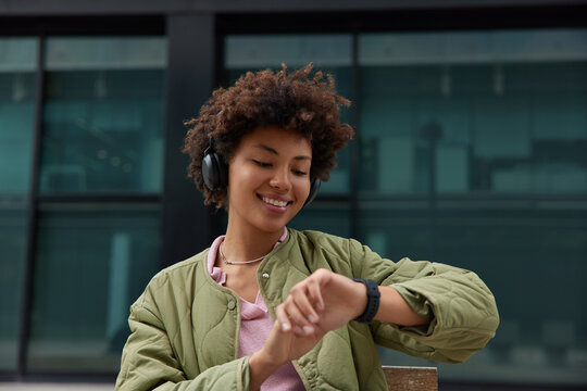 Horizontal shot of good looking Afro American woman with curly hair checks time on smartwatch listens audio track from social media enjoys leisure pastime dressed in anorak. Millennial hipster - Powered by Adobe