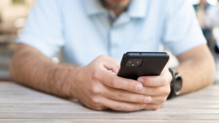 Man chatting outside. Man holding the phone. Black smartphone in hand on wooden table. Man texting in the cafe