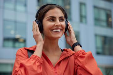 Positive young European woman puts on headphones enjoys favorite music from playlist has outdoor training dressed in sportswear poses against blurred background being full of energy enjoys life