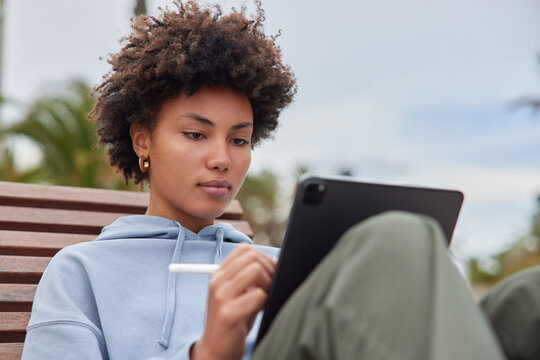 Outdoor shot of beautiful curly haired ethnic woman works on tablet does graphic design draws sketch works as freelance illustrator dressed casually poses outdoor uses electronic pen for writing