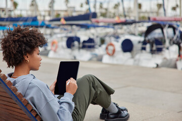 Back view of curly woman works freelance outdoor draws illustration admires view of harbor dressed in casual clothes sits on wooden bench uses electronic pen. Professional photographer edits photos