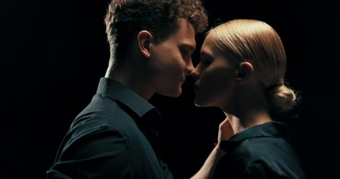 Dark scene studio shot of two people in love beautiful woman with blonde hair stands close to man with shirt unbuttoned who brings lips to woman's forehead, caring for her, safety, moment before kiss