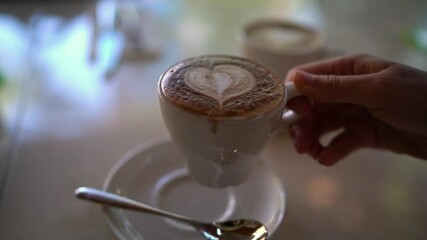 Love for Coffee concept video with latte art heart in Cappuccino. Woman drinking Cappuccino espresso coffee with perfect latte art at trendy cafe. Woman hands taking cup drinking