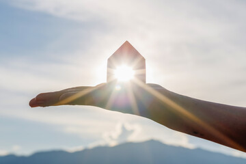 Hand holding wooden house toy against blue sunny sky with sun rays. solar power.