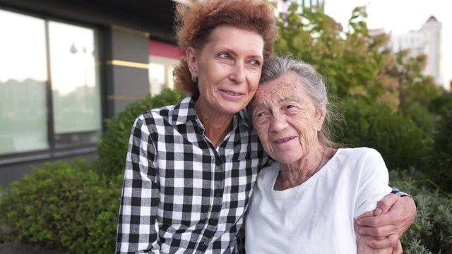 Topic Support And Hanging Out With Old Parents. Caucasian Mature Daughter Expressing Affection Holds And Strokes Hands Of Senior Mother With Deep Wrinkles And Gray Hair While Relaxing On Bench In Park