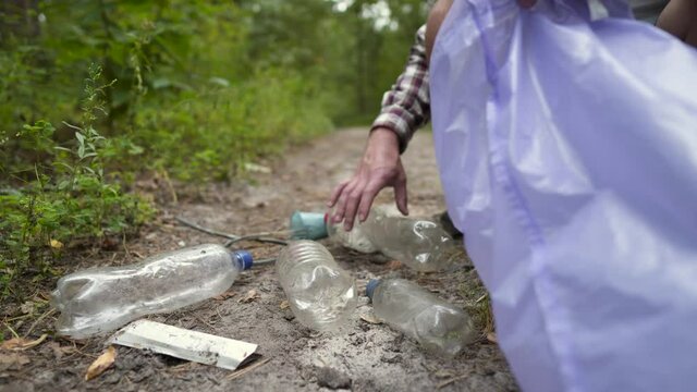 Young male hiker collects rubbish in trash bag while walking along forest path. Trendy sports programs plogging. Volunteer peels off plastic bottles in park and takes them away for recycling.