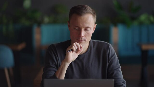 Close-up portrait of ponder young man in office. Designer plans his work and holding a pencil in his hand. Shooting is slow motion from below. Thoughtful serious man sit with laptop thinking solution