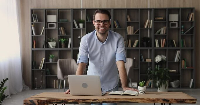 Smiling handsome confident young businessman standing at table with computer in modern office. Happy millennial generation male manager worker employee standing at workplace, successful career concept