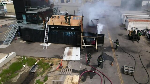 slow motion drone footage of firefighters extinguishing fire on roof and metal container with hoses and full equipment including helmets oxygen masks tanks midday at training facility in California