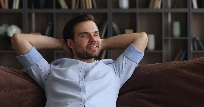 Relaxed happy young handsome man sleeping napping on comfortable sofa with folded hands behind head, sitting on cozy couch in modern living room, visualizing future or meditating alone at home.