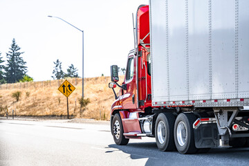 Day cab model red big rig semi truck with roof spoiler transporting cargo in dry van semi trailer running on the highway road with road sign