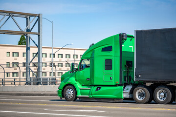 Green professional big rig bonnet semi truck transporting cargo in covered black semi trailer running on the city street bridge