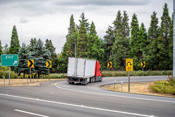 Red big rig semi truck with dry van semi trailer turning on the round highway exit with green trees on the side
