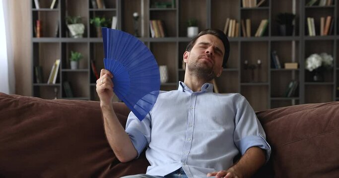 Unhappy stressed young mam using paper fan, cooling fresh air, feeling overheated in living room, suffering from high temperature indoors without conditioning system or hot summertime weather.