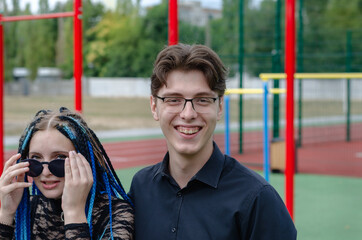 Obraz premium Laughing happy young woman and young man in front of a sports field. Heterosexual couple in love outside. Young adult date. Woman with blue pigtails. Man wearing optical glasses. Love, romance.