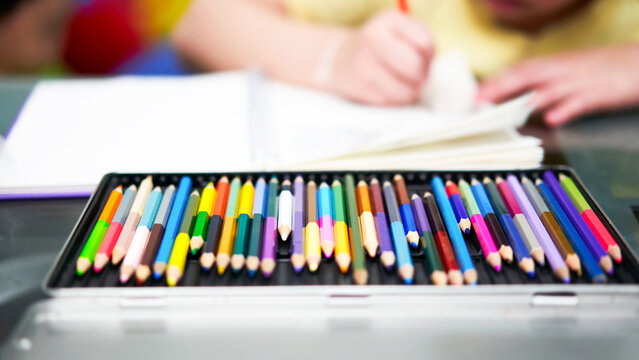 color pencil Crayons of various colors in a palette box, on the table, against the backdrop of a blurry girl drawing with colors.