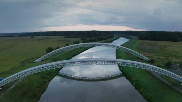 Flying above and below gas pipes across calm river Morava. Aerial sunset footage of two parallel bridges connecting both river banks.