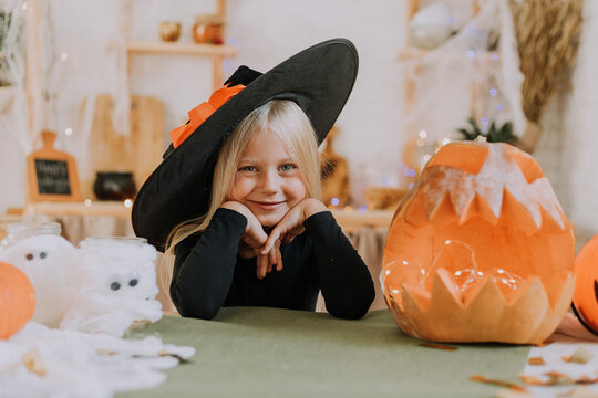 portrait of a little blonde girl with blue eyes, propping her cheeks with her hands, in a witch costume, who is sitting in a room next to a huge pumpkin. halloween concept. High quality photo