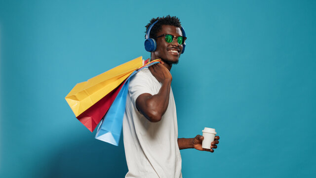 Happy man with handbags dancing after shopping spree. Cheerful person with sunglasses listening to music on headphones while holding bags with clothing for sale purchase on black friday - Powered by Adobe