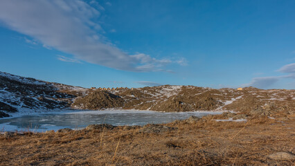 A small frozen lake is surrounded by hills. Snow on the ice and on the ground. In the distance, against the blue sky - the houses of the tourist base. Beautiful clouds. Siberia. Olkhon Island