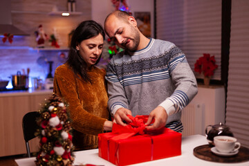 Cheerful family opening xmas decorated gift with ribbon on it during christmas holiday standing in x-mas kitchen. Happy couple married celebrating winter season. Santa-claus tradition