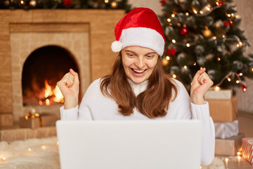 Extremely happy woman working on laptop near Christmas tree and fireplace, wearing santa claus hat and white sweater, clenching fists with success, posing in living room with x-mas decorations.