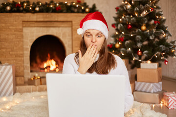 Fototapeta na wymiar Indoor shot of surprised attractive girl working in eve of new year with laptop, posing in living room near fireplace and x-mas tree, looking at pc with shocked expression, covering mouth with palm.