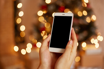 Closeup shot of woman hand holding phone with black blank screen for advertisement or promotional text with christmas tree on background.