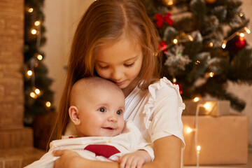 Fototapeta na wymiar Indoor shot of two sisters posing near Christmas tree at home, elder girl hugging infant baby with love, merry Christmas and happy new year, children in festive room.