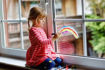 Adoralbe little toddler girl with rainbow painted with colorful window color during pandemic...
