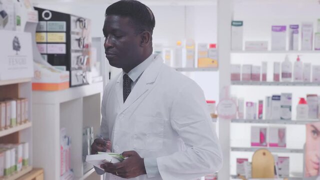African-american male pharmacist standing with notebook in hands in salesroom of drugstore. High quality 4k footage