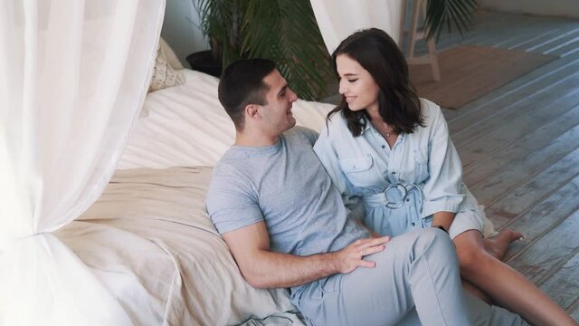 Slow motion portrait of happy young couple sitting in front of bed and hugging. Attractive woman kissing her boyfriend and laughing. Romantic and love concept. High quality FullHD footage