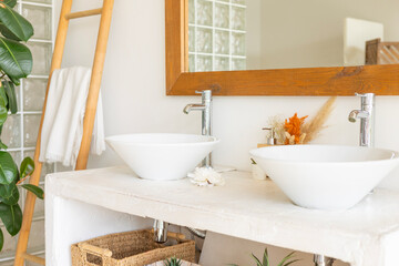 stylish and modern design bathroom interior with two white sinks, wicker baskets for cosmetics, mirror in wooden frame, dried flowers in vases, home plants in pot in Balinese style with white walls